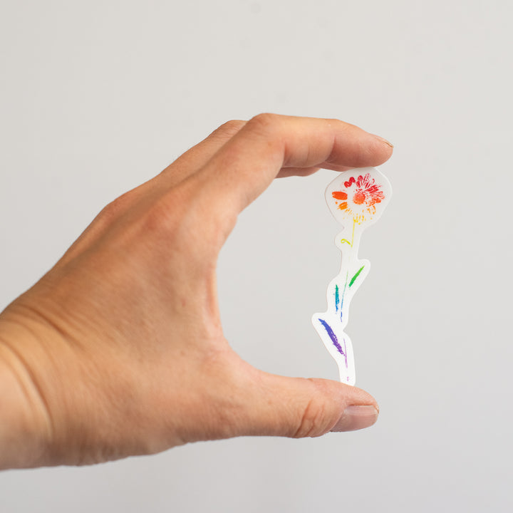 Hand holding a small, colorful daisy sticker against a plain background