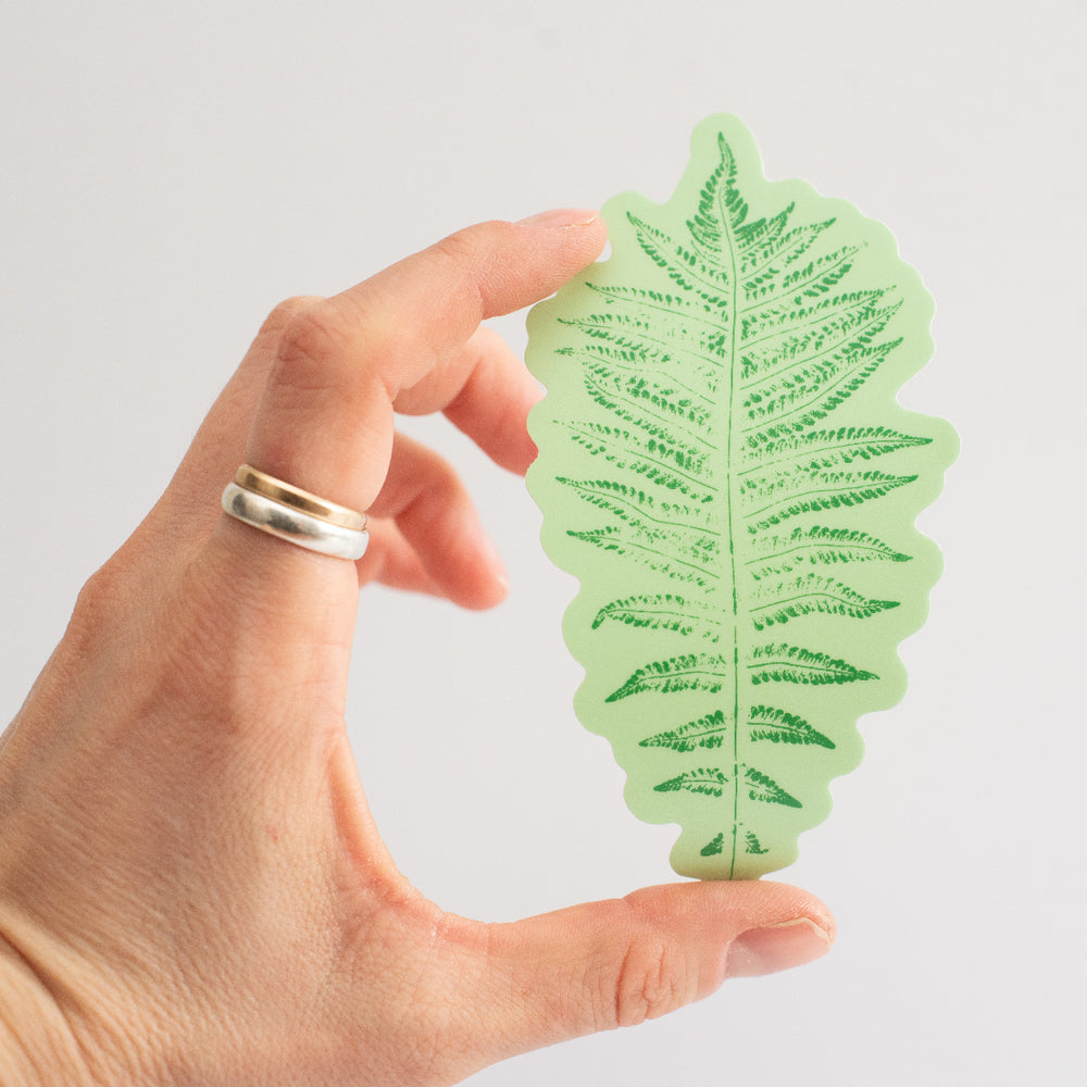Hand holding a green fern leaf-shaped sticker against a light background