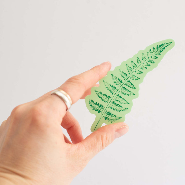 Hand holding a green fern leaf-shaped sticker with detailed patterns on a light gray background
