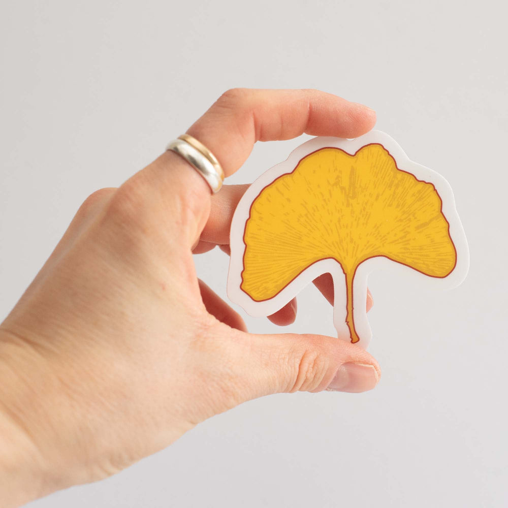 Hand holding a yellow ginkgo leaf-shaped sticker against a light gray background