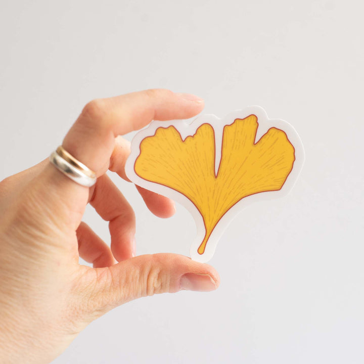 Hand holding a yellow ginkgo leaf-shaped sticker against a light gray background