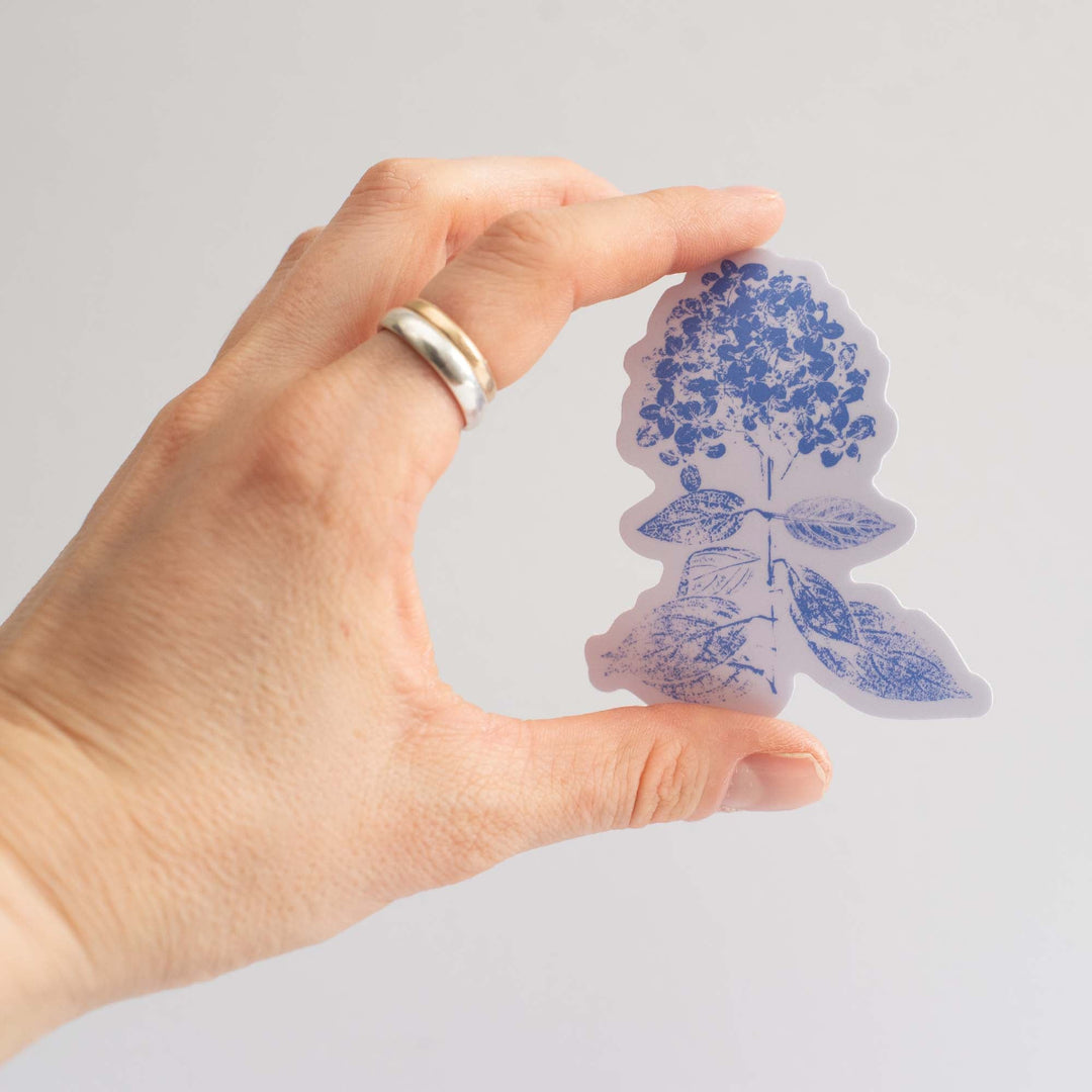 Hand holding a two-tone blue hydrangea floral sticker against a gray background