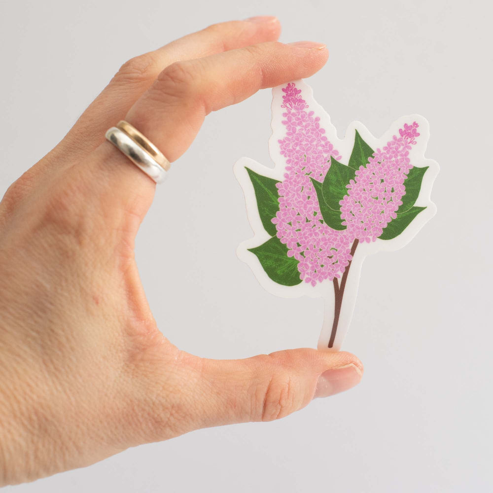 Hand holding a sticker of lilacs and green leaves against a light gray background