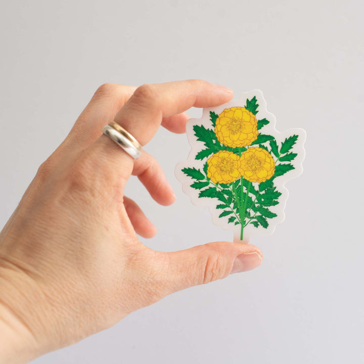 Hand holding a sticker with marigold yellow flowers and green leaves in front of a light gray background