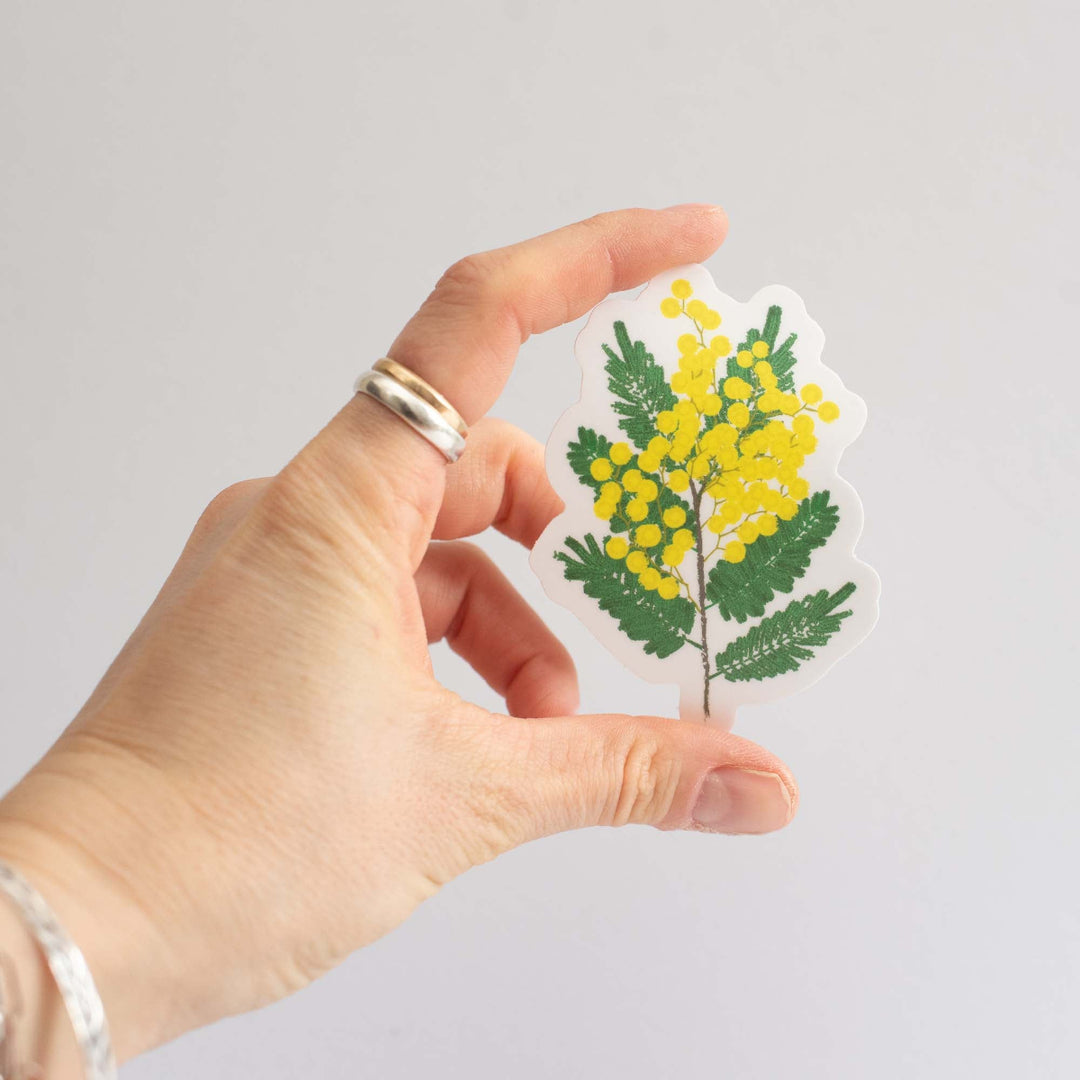 Hand holding a sticker with yellow flowers and green leaves on a plain background