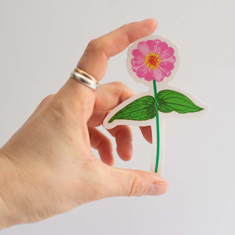 Hand holding a colorful pink zinnia flower sticker against a light gray background