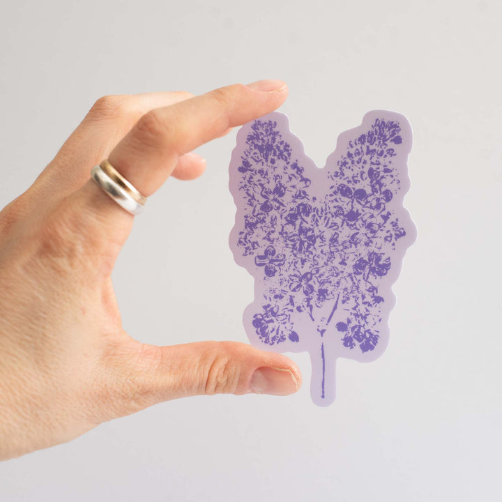 Hand holding a purple lilac flower sticker against a light gray background