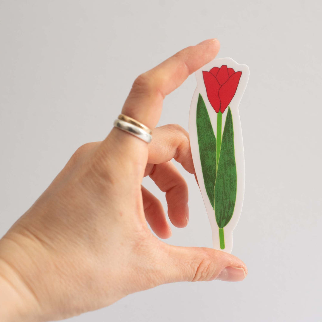 Hand holding a cutout sticker of a red tulip with green leaves against a light gray background