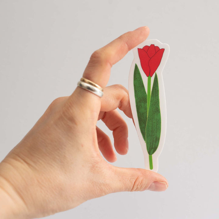 Hand holding a cutout sticker of a red tulip with green leaves against a light gray background