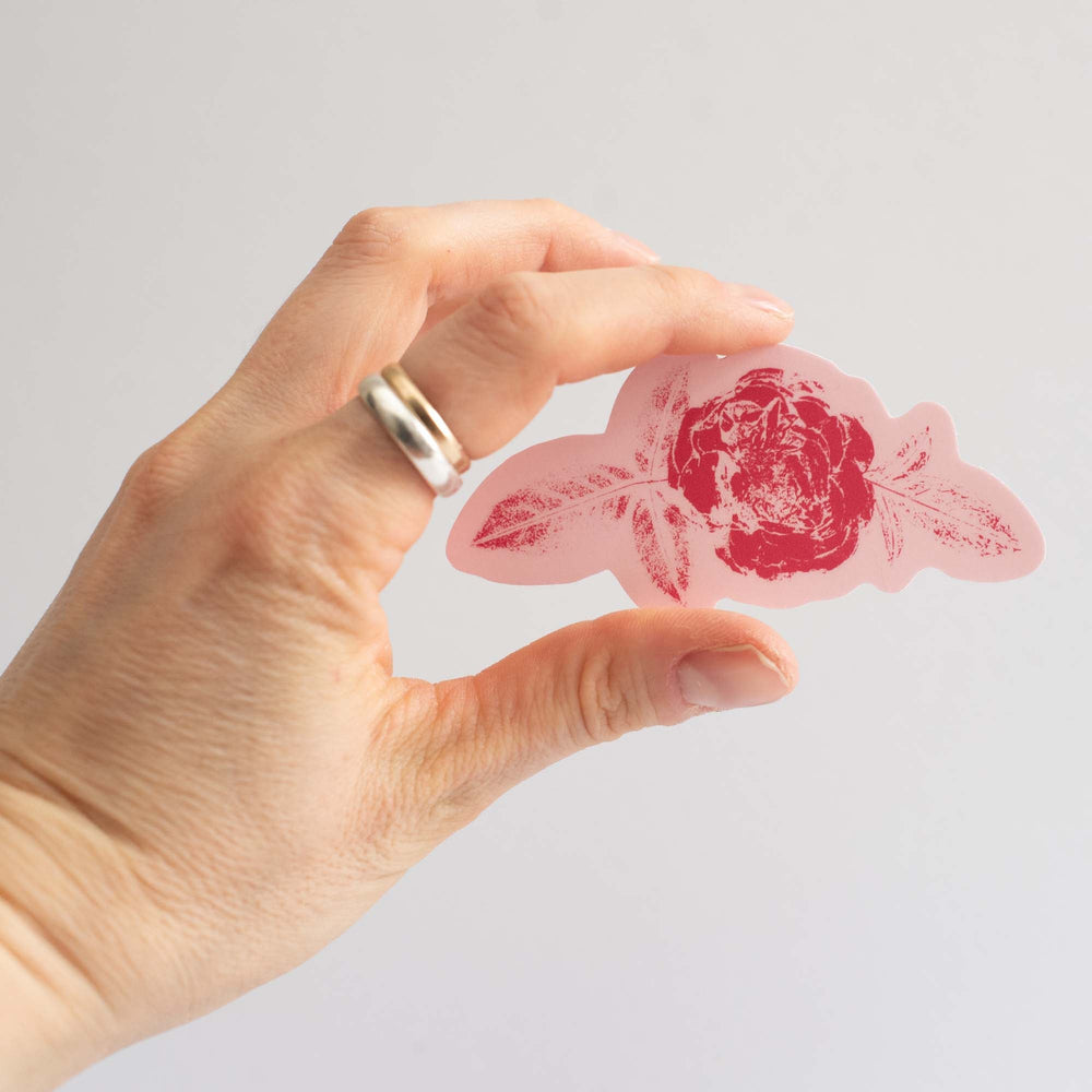Hand holding a two-tone pink rose sticker  against a light gray background