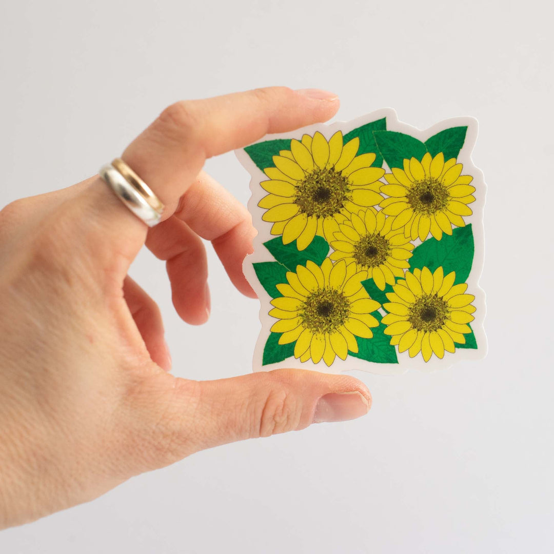 Hand holding a square sticker with sunflower design in front of a light gray background