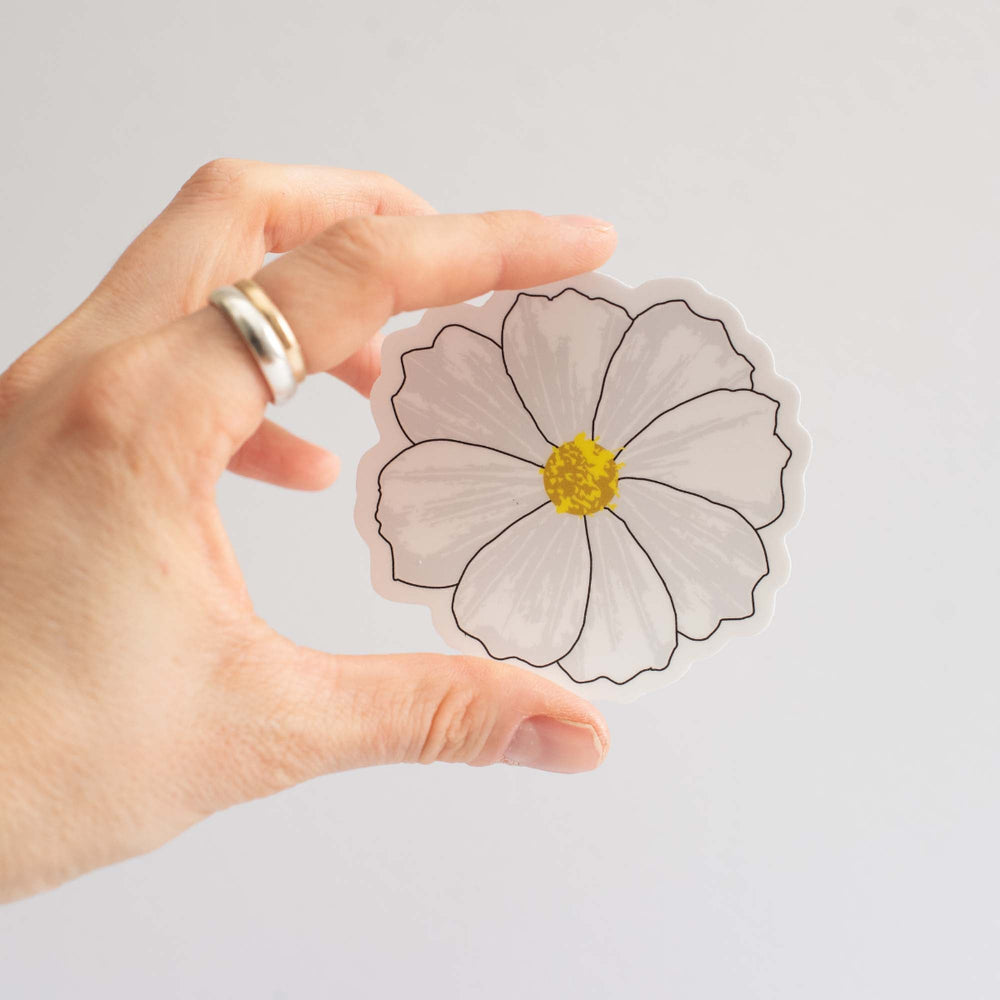 Hand holding a white cosmos flower-shaped sticker against a light gray background